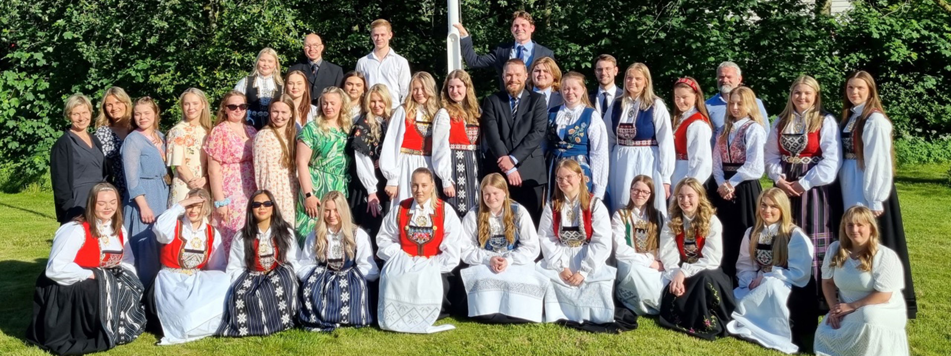 Eit gruppebilde av elevar og lærarar på grasplenen utanfor skulen, alle er pynta med dress, kjolar og bunad.