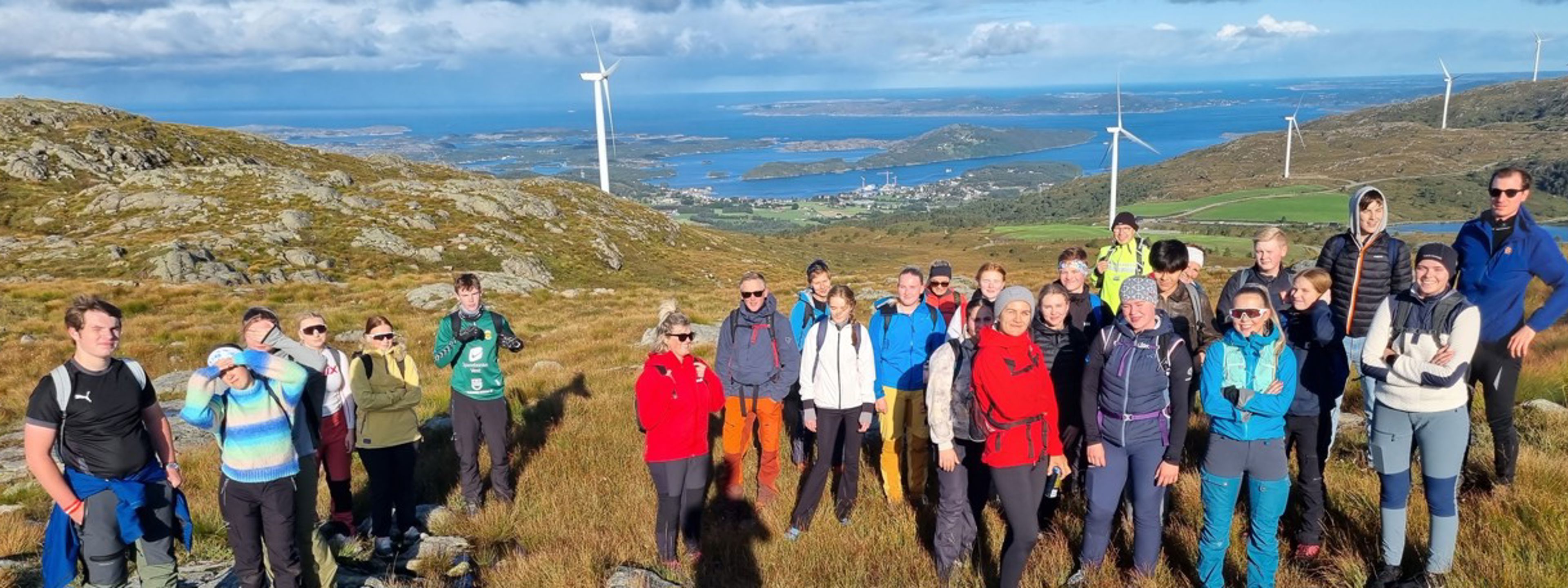 Mange elevar og lærarar på ein fjelltopp med ryggen mot vindmøller