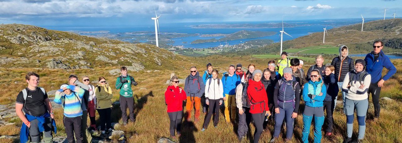 Mange elevar og lærarar på ein fjelltopp med ryggen mot vindmøller