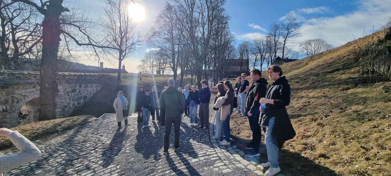 Mange elevar som står på brustein på toppen av Akershus festning og høyrer på ein lærar som står med ryggen til. Sol og blå himmer