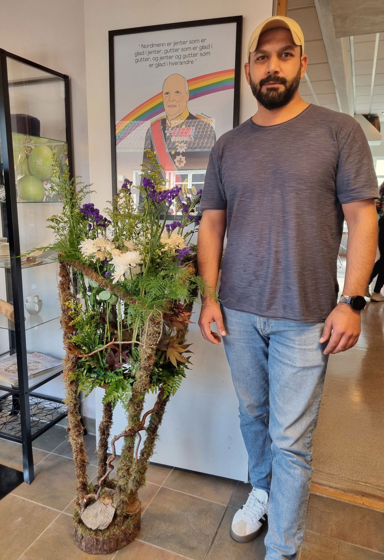 Ein mann med caps som står framfor ein stor blomsterbukett i ei mosekledd ramme