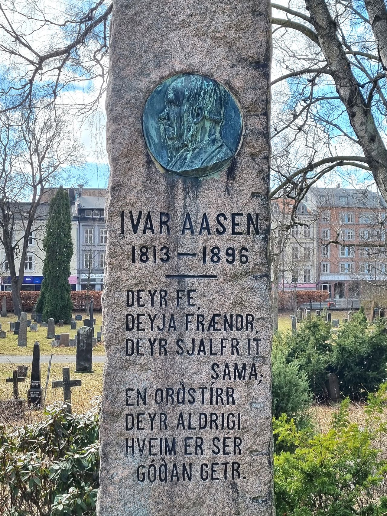 Eit foto av ei gravstøtte der det står IVAR AASEN 1813-1896, samt eit vers på norrønt og ei metallplate med eit portrett av Ivar Aasen