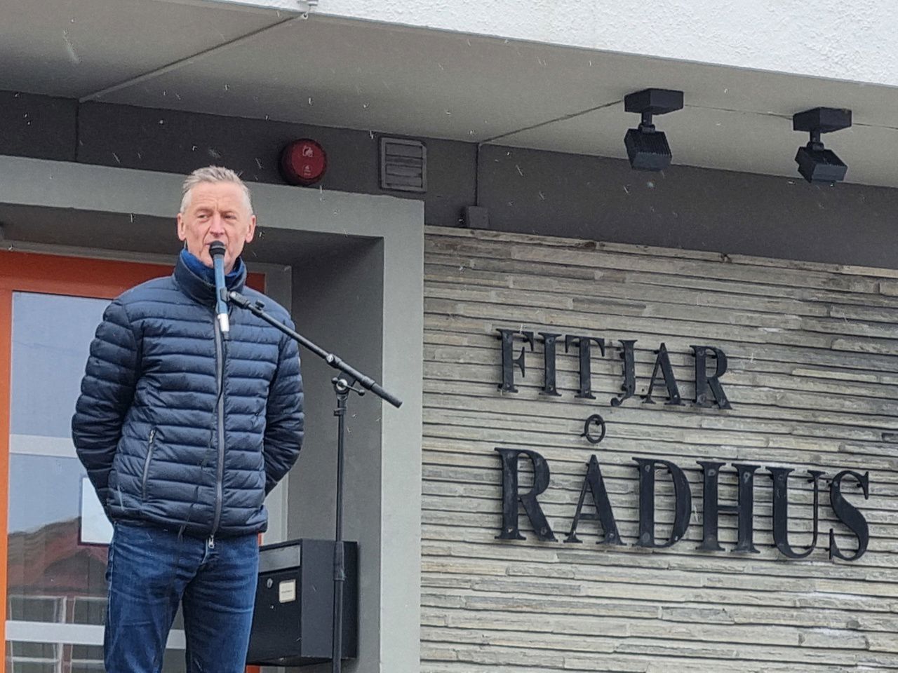 Ein mann i blå boblejakke som held ein tale framfor ein mikrofon. I bakgrunnen står det Fitjar rådhus på ein vegg