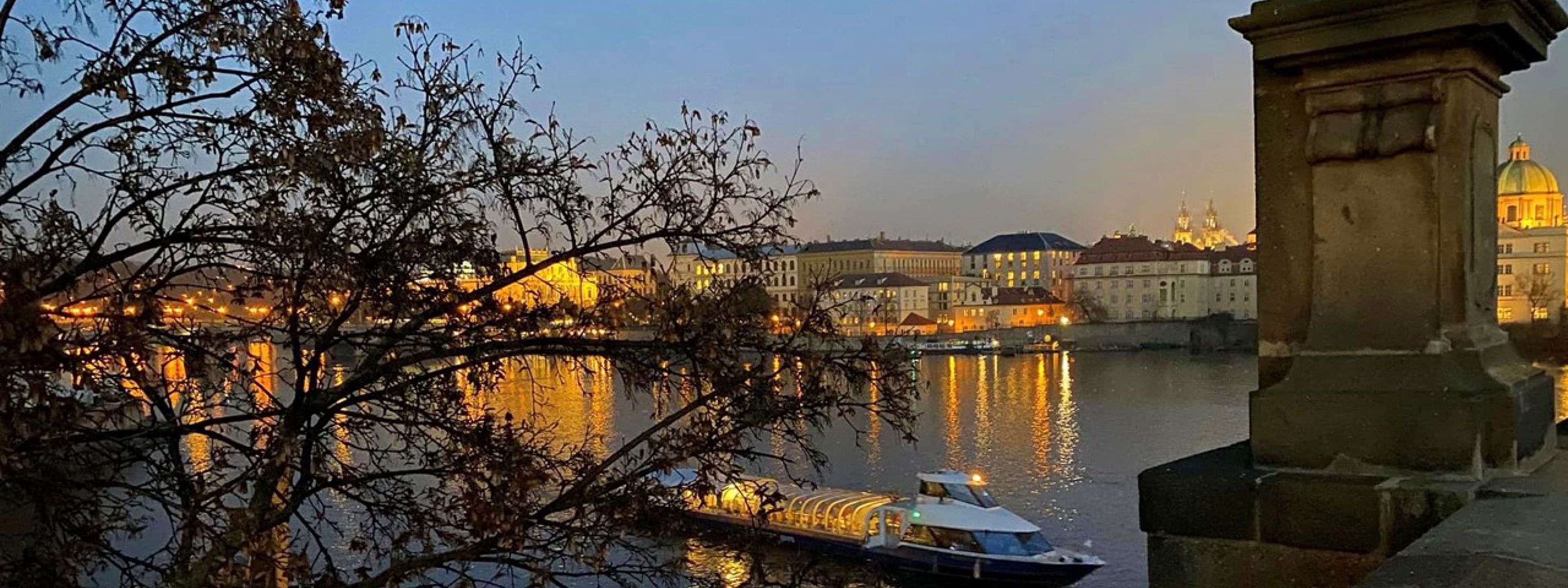 Ei elv i Praha i skumringen, på elva er det ein båt, i framgrunnen er det ein statue