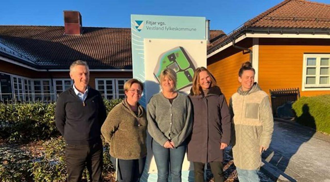 Fem mennesker som står framfor eit skilt som det står Fitjar vidaregåande skule på, i bakgrunnen ser me skulen