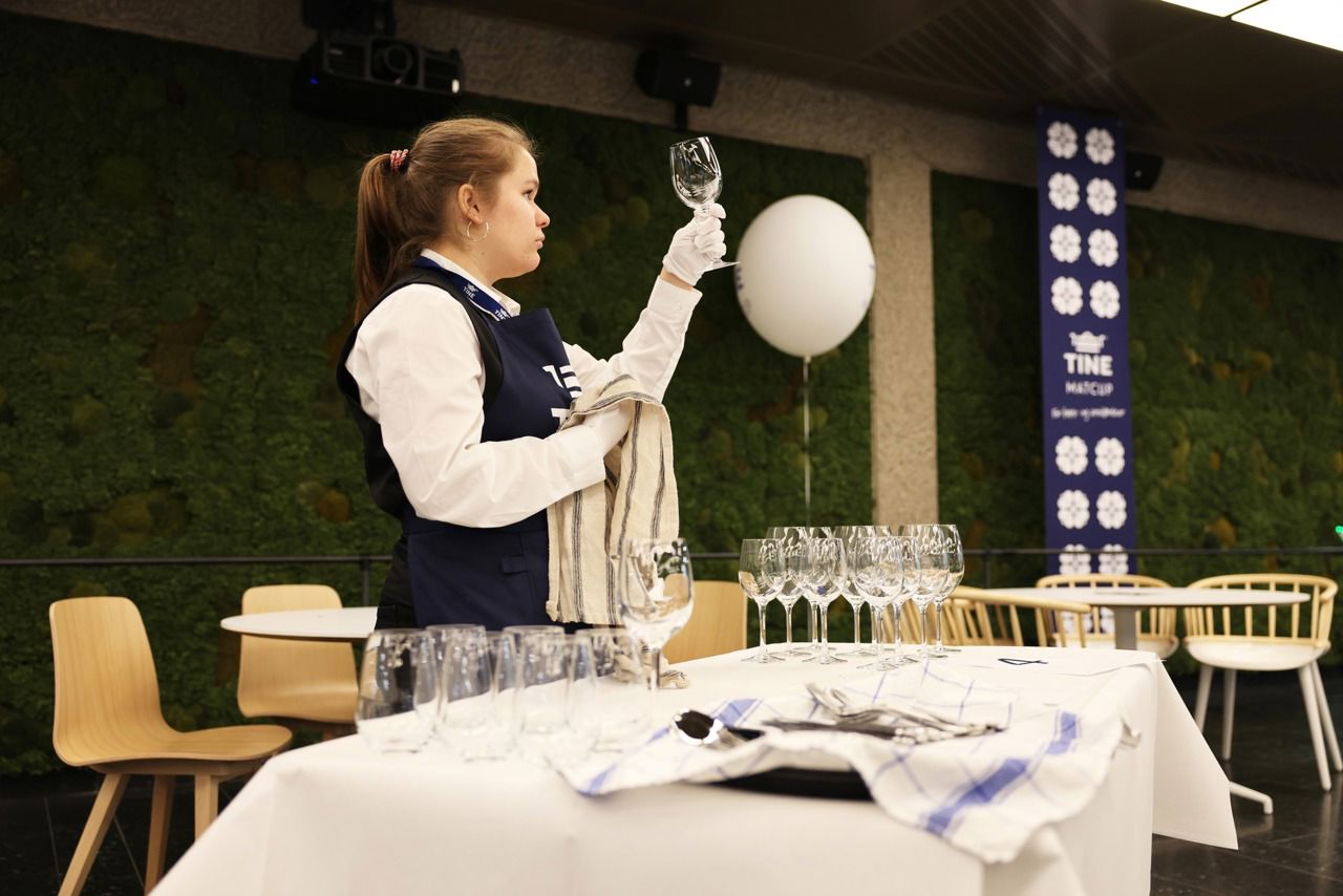 Ein elev i servitøruniform som pusser eit glass framfor eit dekka bord