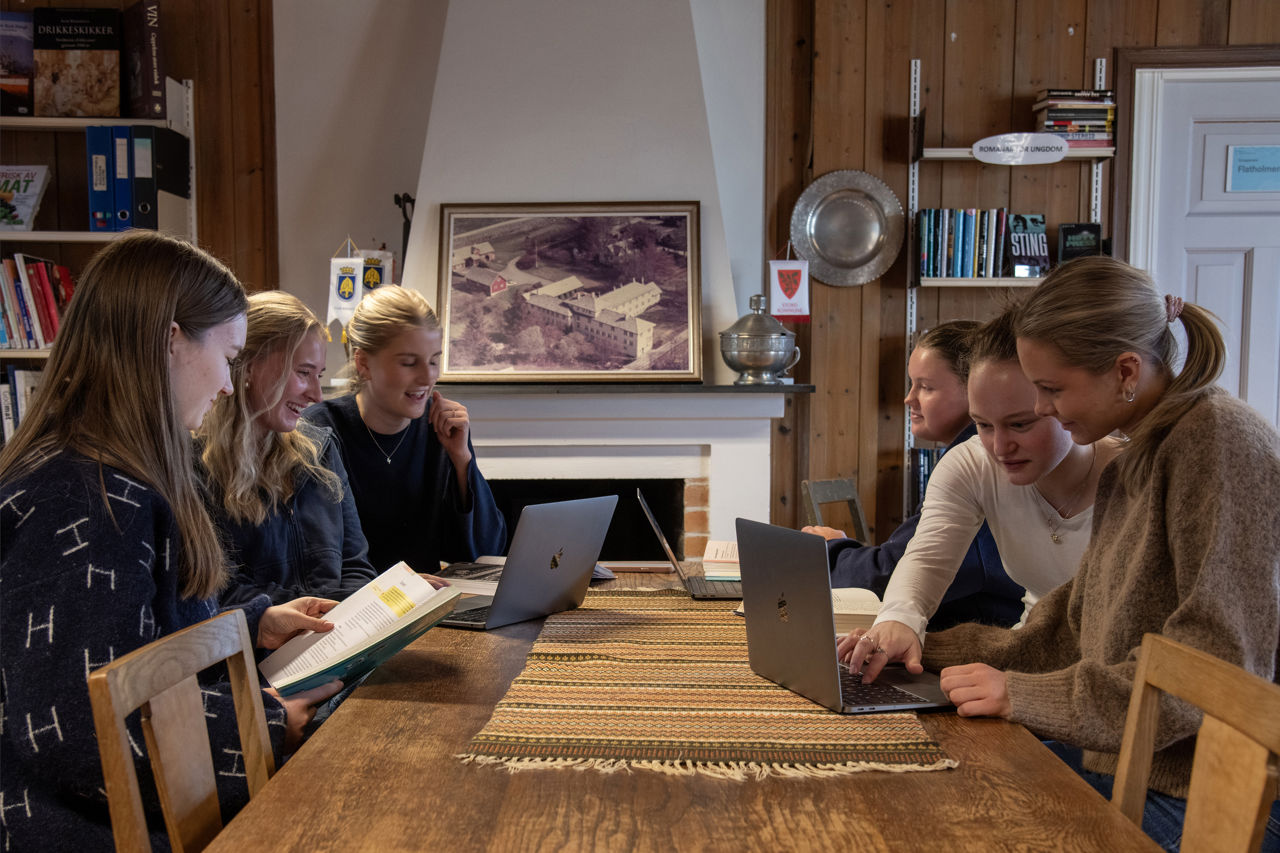 Seks elevar, jenter, sit ved eit langbord på biblioteket og arbeider med PC og bøker