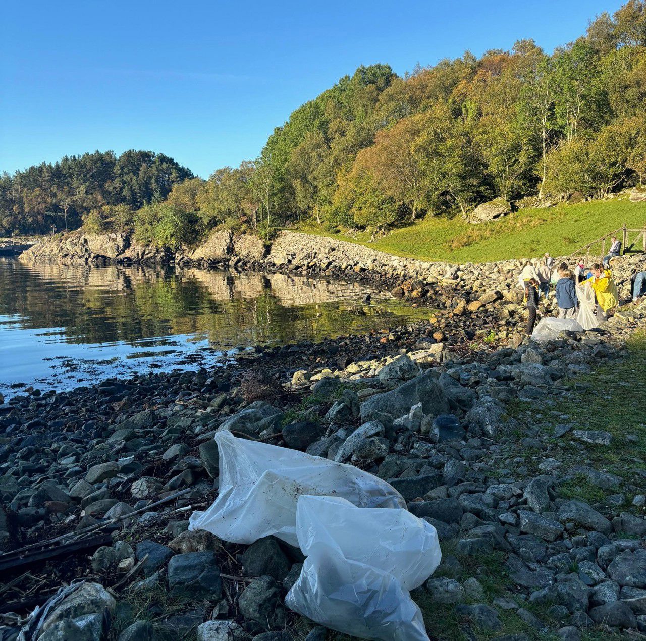 To søppelsekkar i framgrunnen, ei strand i bakgrunnen, og så grøne bakkar og trær. Me ser nokre elevar som ryddar søppel langt unna