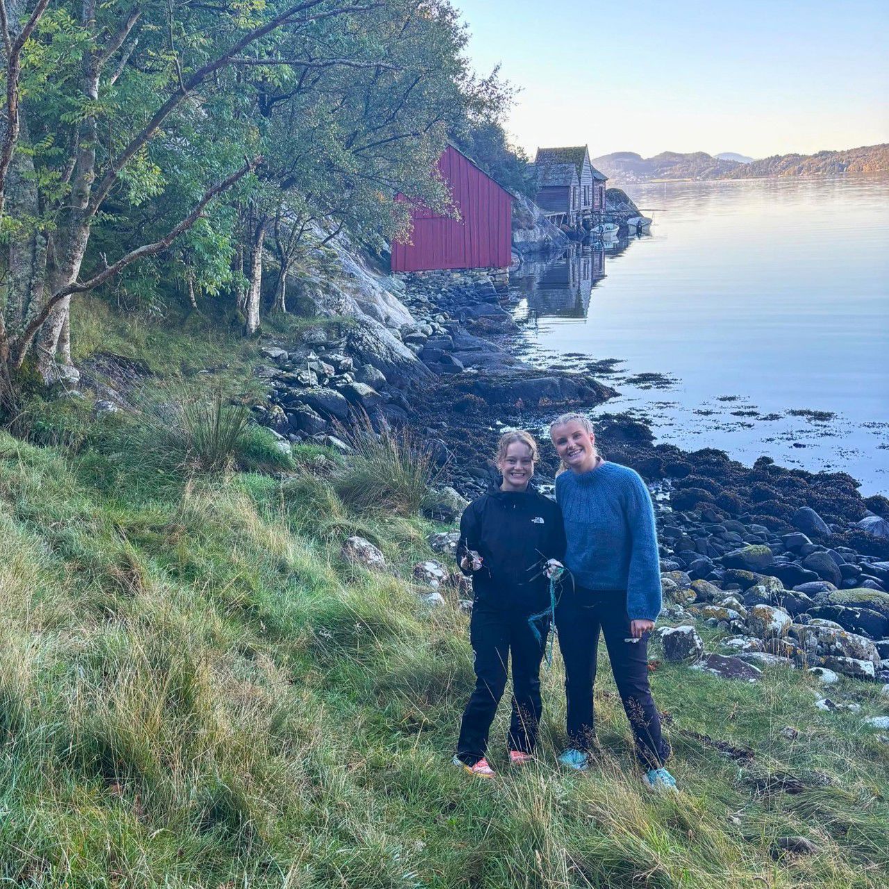 To elevar som står i ei strand med søppel i hendene, i bakgrunnen naust, skog og fjord