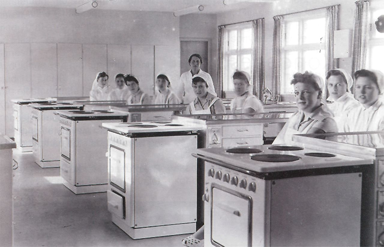 Svart kvitt bilete frå 1950-talet der husmorelevane sit i klasserommet attmed komfyrane