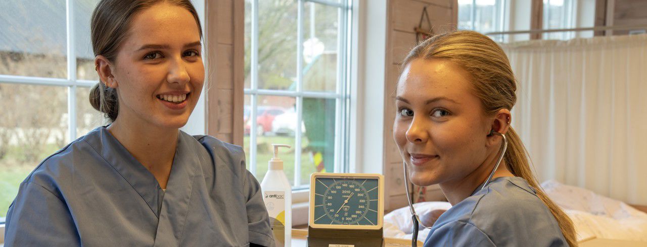 To elevar i framgrunnne, på kvar si side av biletet. Den eine har helseuniform på.  I bakgrunnen sit det to elevar i kvite frakkar som måler blodsukker