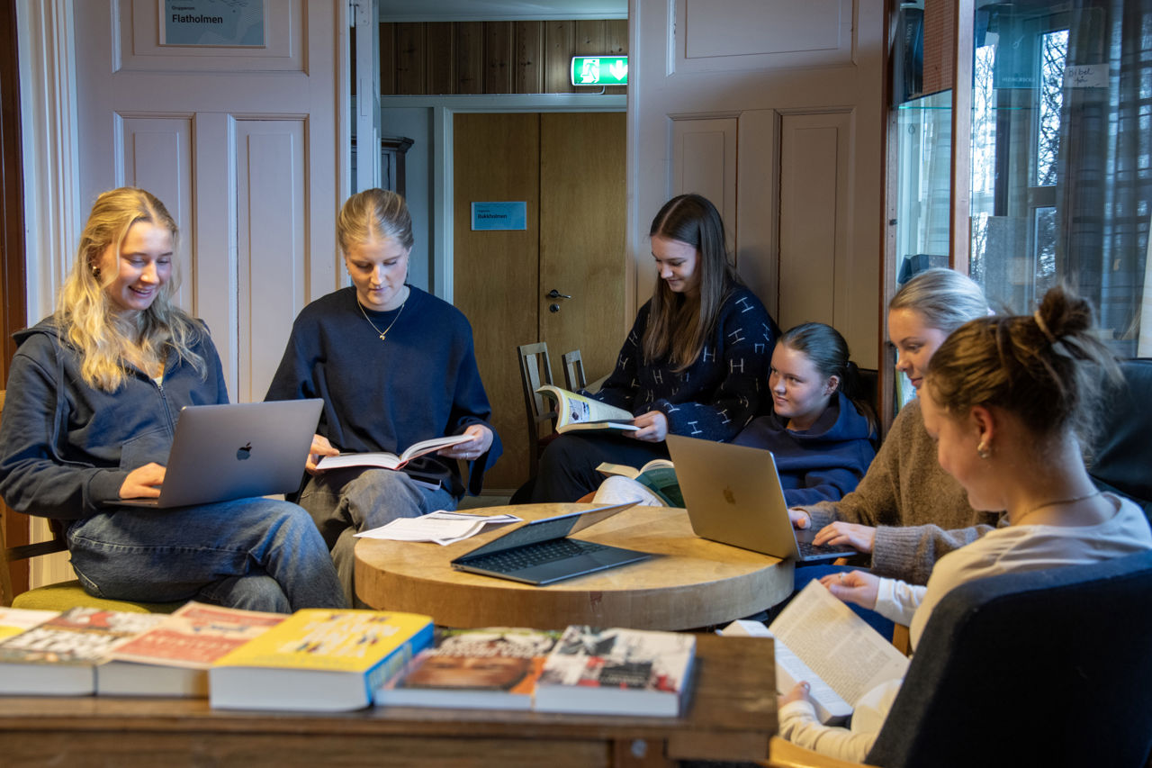 Seks elevar, jenter, som sit rundt eit rundt bord i kantina og arbeider med bøker