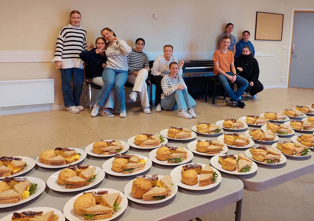 Eit langbord med mange tallerkar med mat og i bakgrunnen står mange elevar