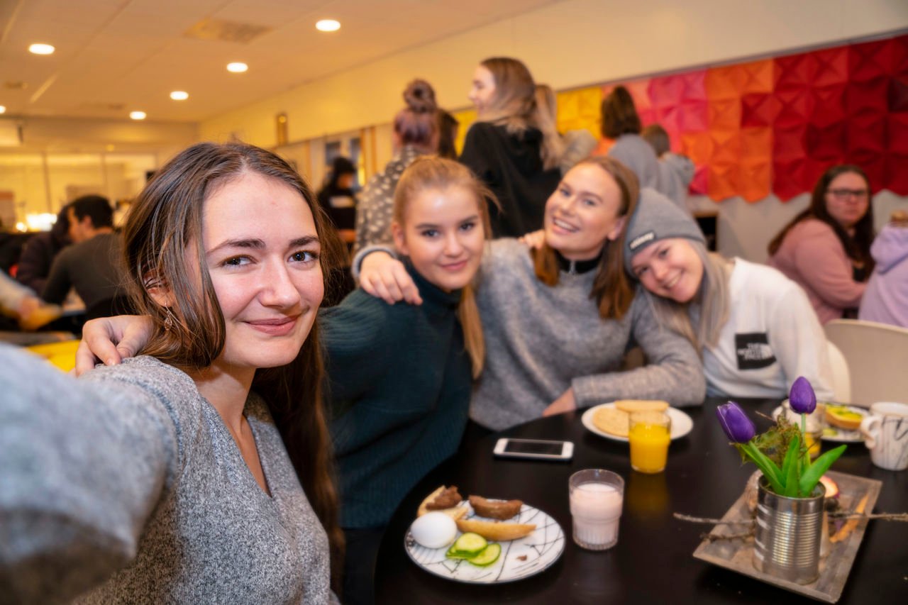 Fire jenter som sit i kantina og held rundt kvarandre og smiler, mat står på bordet.