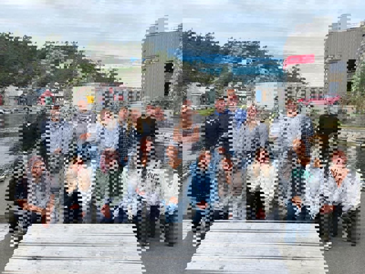 Ei gruppe elevar og tre vaksne som står framfor vannkanten. Hytter og naust i bakgrunnen